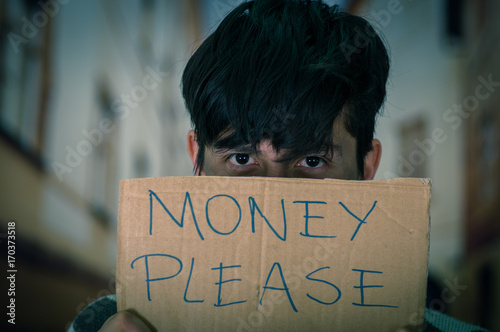 Portrait of a homeless with cardboard description of money please, hiding hald of his face, in a blurred background photo