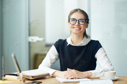 Young smiling businesswoman in eyeglasses sitting by her workplace