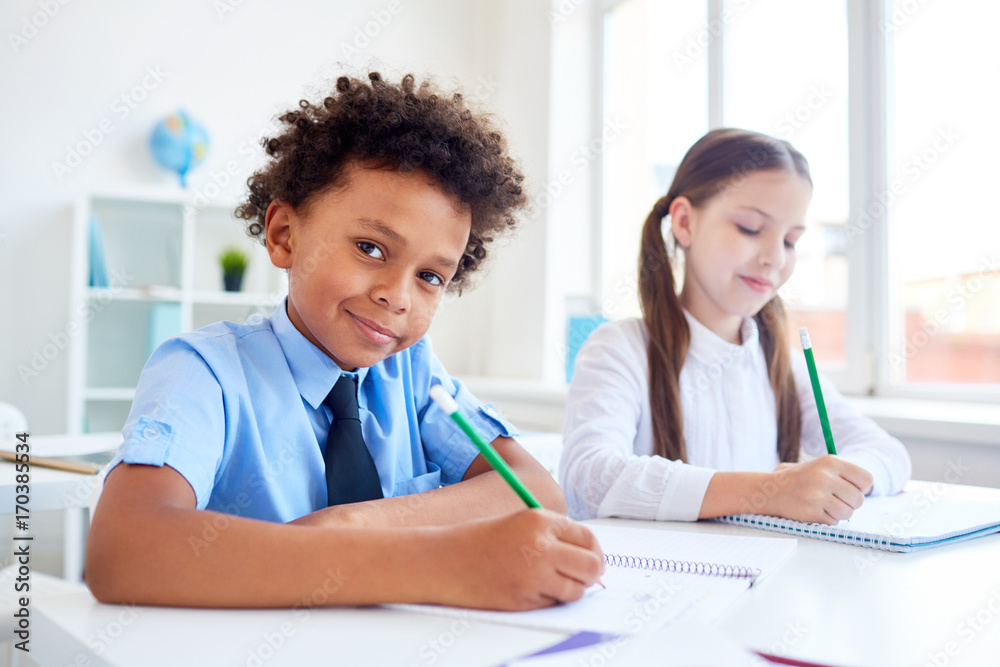Cute boy of African ethnicity drawing with pencil in copybook with classmate on background Cute boy of African ethnicity drawing with pencil in copybook with classmate on background