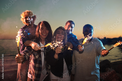 Cheerful friends with sparklers celebrating holiday by seaside