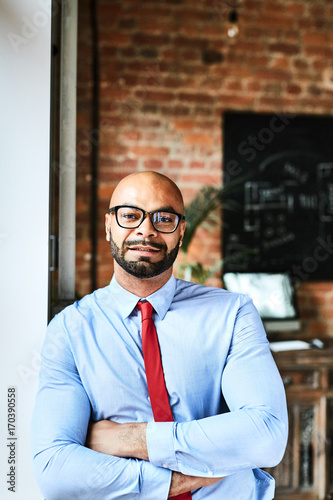 Mixed race businessman in eyeglasses posing for photography with arms crossed while  leaning to wall, interior of modern office on background