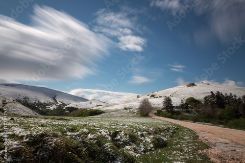 Long exposure photo of a mountain scenery with green grass and melting snow near a road, under a blue sky with moving clouds