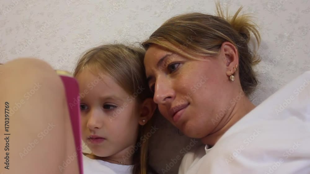 Charming little girl and her beautiful young mom are using a digital tablet and smiling while sitting on sofa at home Charming little girl and her beautiful young mom are using a digital tablet and smiling while sitting on sofa at home