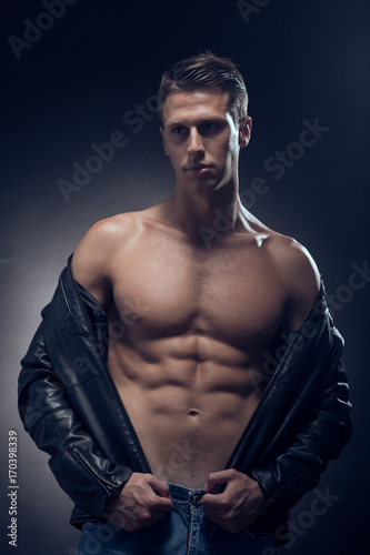 fitness model posing, muscular abs chest, upper body shot, one young adult Caucasian man, black background, studio, jeans, leather jacket