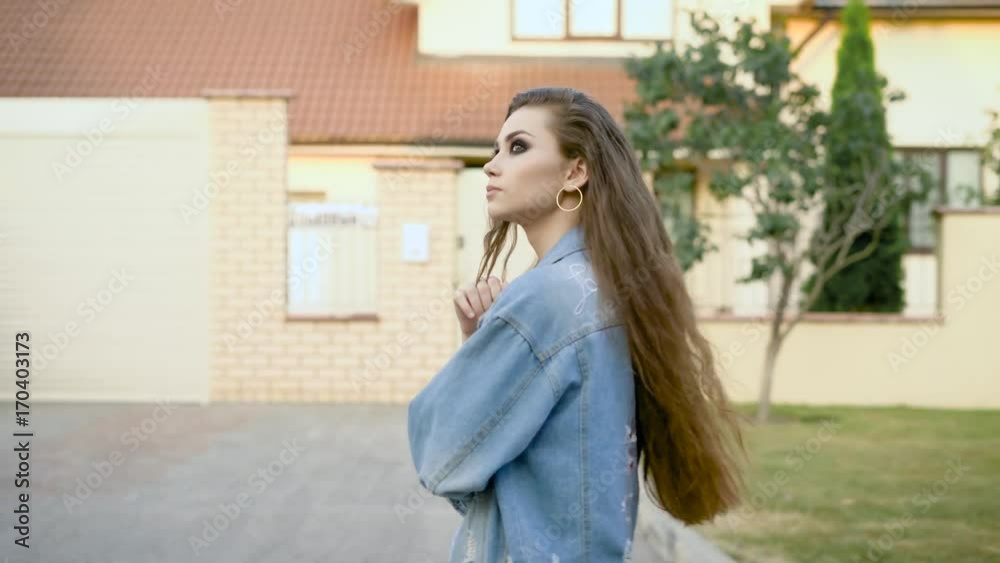 thoughtful young woman with long dark wavy hair is walking over yard along cottages in summer evening, side view thoughtful young woman with long dark wavy hair is walking over yard along cottages in summer evening, side view