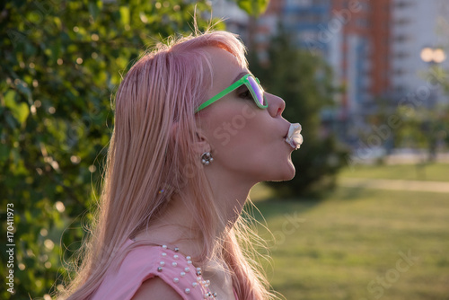 Young girl in green glasses with long hair puffs bubble of chewing gum in summer