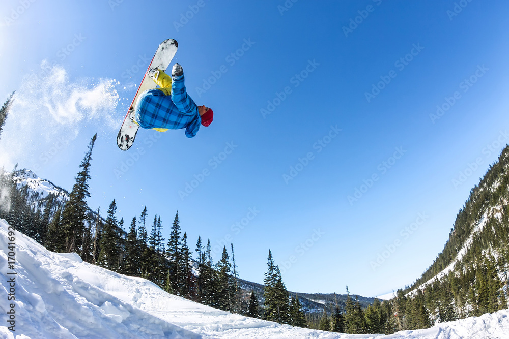 Snowboarder freerider jumping from a snow ramp in the sun on a background of forest and mountains