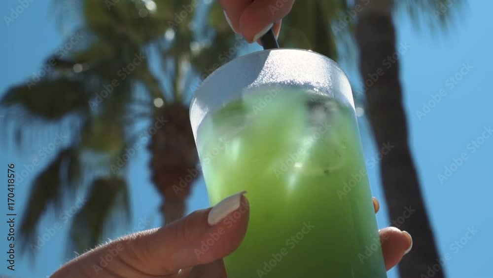 Unrecognizable woman holding green refreshing drink with ice cubes and stirring it with straw.