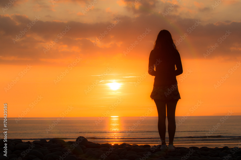 Young woman stands looking out to sea at the sunset