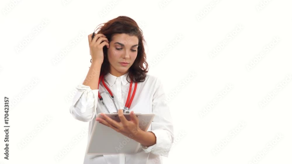 Friendly young female doctor reviews and writes notes on a clipboard. Portrait of young medical professional with stethoscope and lab coat isolated on white background Friendly young female doctor reviews and writes notes on a clipboard. Portrait of young medical professional with stethoscope and lab coat isolated on white background