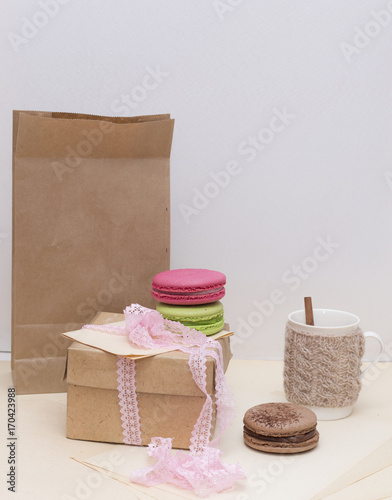 Beige knitted mug and colorful macaroons on rough paper bag background. Kraft box and pink laces. Nude still life.