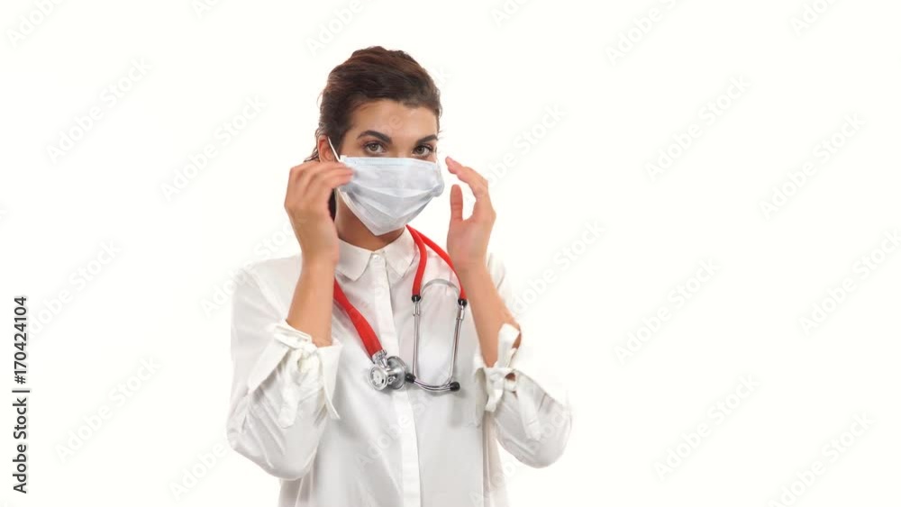 Portrait of a female doctor looking in camera and taking on a protection mask. Close up of a female surgeon with stethoscope and lab coat isolated on white background dressing up surgical mask Portrait of a female doctor looking in camera and taking on a protection mask. Close up of a female surgeon with stethoscope and lab coat isolated on white background dressing up surgical mask