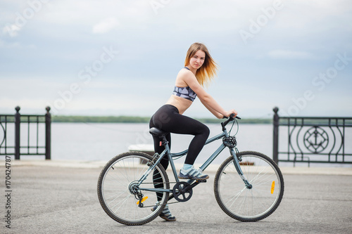 Smiling young female standing with bicycle on embankment and looking at camera. Girl dressed in a sports top and leggings.