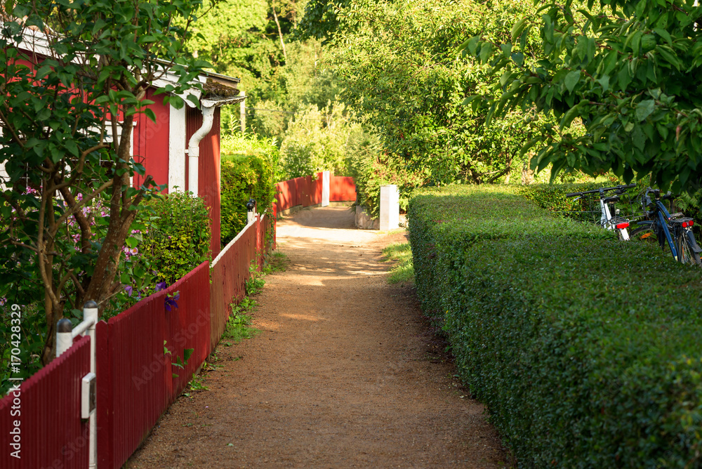 Obraz premium Narrow gravel lane in allotment area. Red and white cottage on one side and green hedge on the other.