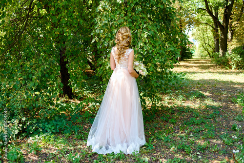Beautiful bride in long wedding dress with bridal bouquet of flowers walking in the park outdoor. Happy bride in wedding dress on summer day, free space