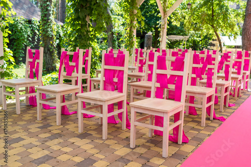 Wedding chairs on each side of archway with pink cloth. Place for wedding ceremony decorated in pink color, wooden chairs for guests outdoors. Wedding ceremony in pink color