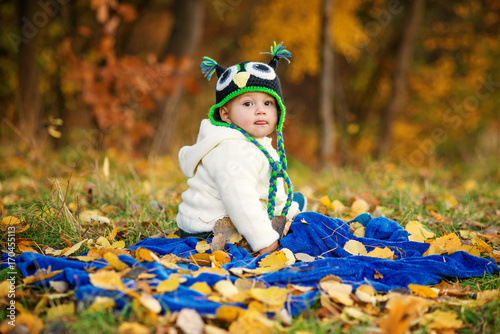 Little happy boy in warm clothes shows a tongue  sitting on a blue plaid and playing with toys on a green lawn with autumn leaves on the ground.