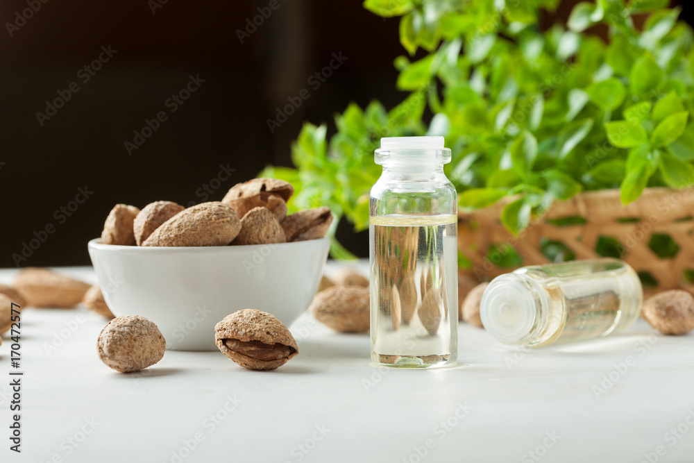 Small glass bottle with oil in it and white bowl full of almond in shell on a white table with greenery in wicker basket