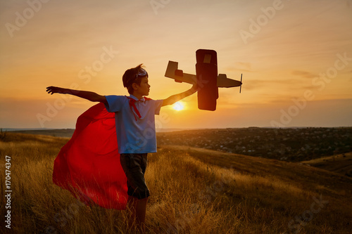 A happy boy in a superhero costume is playing with an airplane on the nature at sunset.