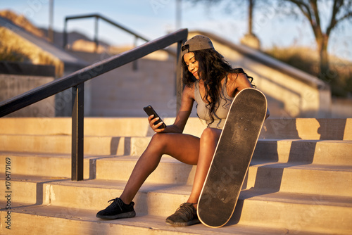 young african american woman with skateboard using smart phone at park while resting on steps
