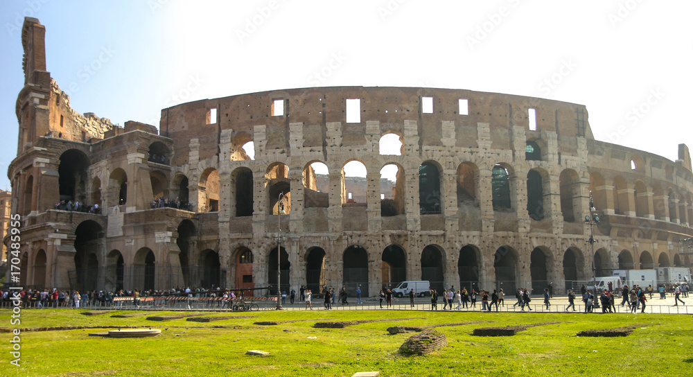 Naklejka premium Wide exterior of the Coliseum in Rome with clear morning skies and bright green grass, backlit by a lens flare