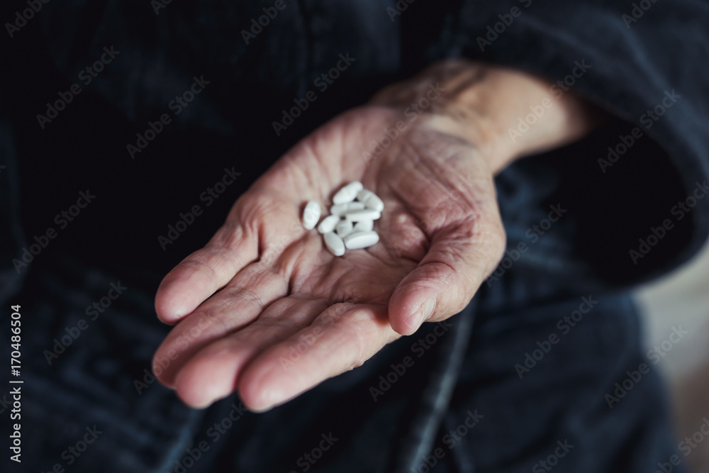 Old woman shows in hand handful of white tablets or pills on dark background