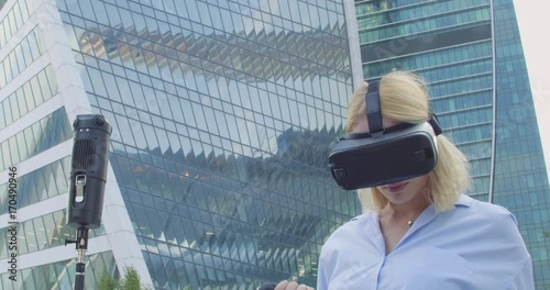 A blond woman in a blue shirt with a VR glasses and camera 360. Skyscrapers are on the background 4K