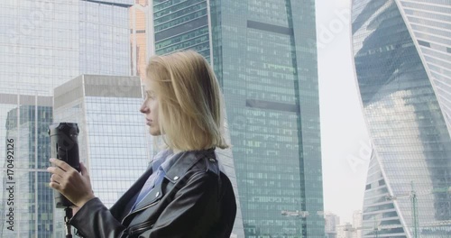 A woman in a blue shirt and black leather jacket takes on a 360 VR camera. Big city and skyscrapers at the background