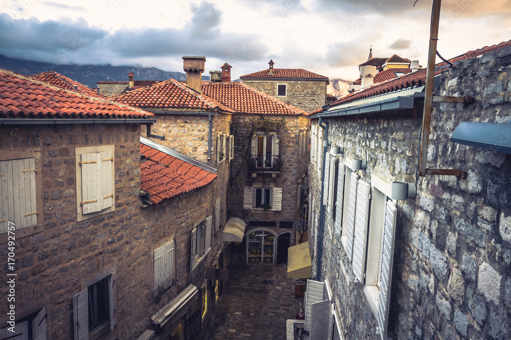 Vintage european city street  with orange tile roofs and ancient building facade in front of dramatic sunset sky with antique architecture in old European town Budva in Montenegro 