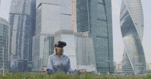 A woman in a blue shirt and VR glasses is sitting in a lotus pose. Skyscrapers are on background