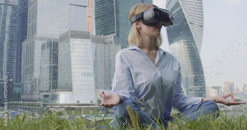A woman in a blue shirt and VR glasses is sitting in a lotus pose. Skyscrapers are on background