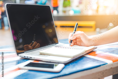 A woman's hand working on a notebook computer and writing on a notepad with a pen on a wooden desk.