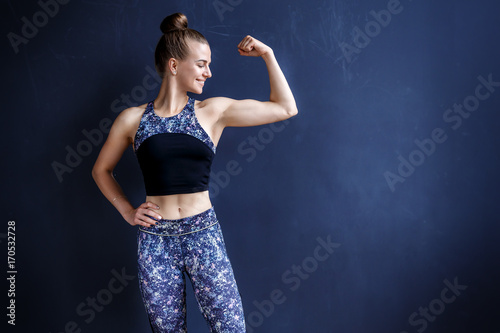 Beautiful fitness model is posing in front of the dark wall in a dark training suit