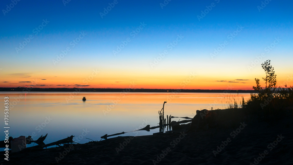 The dark silhouette of two lovers kissing in the water at night in the river on the horizon against a beautiful orange sunset sky at the shore with beach sand and the water tank at the camp.