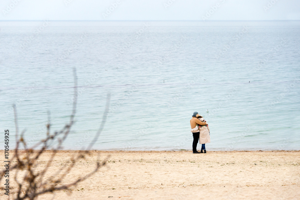 Happy loving couple embracing wearing warm clothes standing on ocean coast. Cold season, sea shore, empty beach in autumn or winter. Happy loving couple embracing wearing warm clothes standing on ocean coast. Cold season, sea shore, empty beach in autumn or winter.