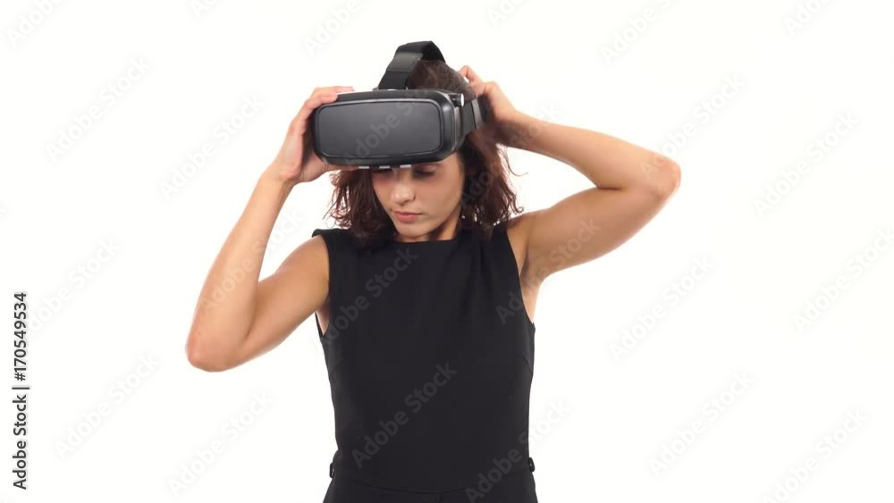 Attractive young woman in black dress taking on, looking around then taking off virtual reality glasses isolated over white Attractive young woman in black dress taking on, looking around then taking off virtual reality glasses isolated over white