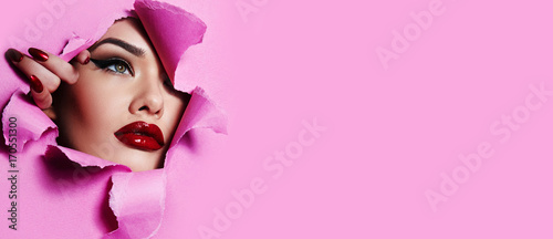 the face of a young beautiful girl with a bright make-up and with plump red lips peeks into a hole in pink paper. 