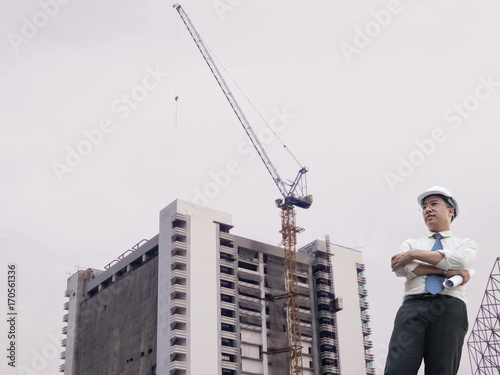 Young businessman/ engineer male crossed arms with blueprint in hand ,he wear white safety helmet ,background is blurred background of industrial construction site