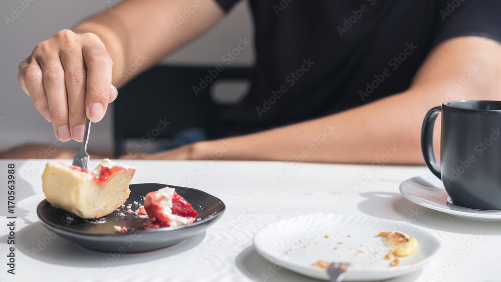 Closeup image of a man using fork to cutting sweet dessert in a plate with black cup of coffee on white vintage wooden table in cafe