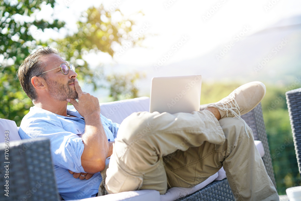 Smiling mature man using laptop, relaxing in outdoor sofa
