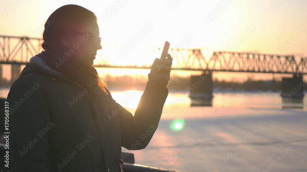 Fototapeta premium Young attractive girl is walking along the waterfront on the background of a railway bridge and a beautiful sunset, holding a phone in hands, writes a message.