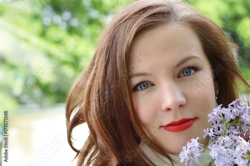 Beautiful face of a young red-haired girl with white skin, blue eyes and red lipstick on her lips. A branch of lilac.