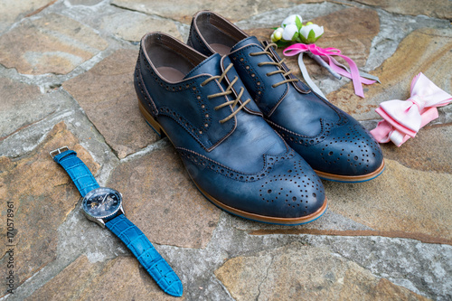 Blue leather groom shoes, watches, boutonniere and pink bowtie on brown natural stone. Groom wedding accessories. Man watches, bow-tie, boutonniere and footwear on stone background