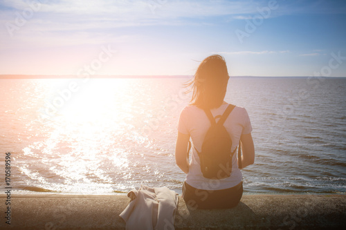 Fotomurale Girl sits on the stone embankment of coast sea and looks at the sun reflects on the surface of the water