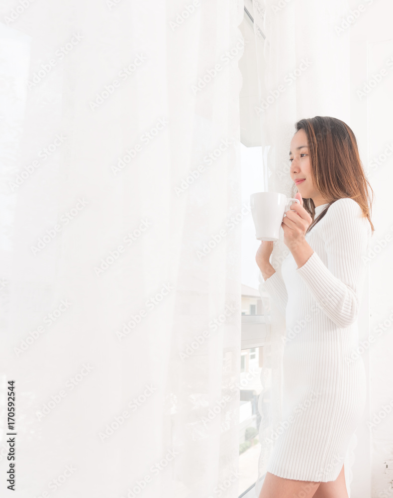 Beautiful asian young woman is looking out the window and holding a cup of coffee in the morning time. Beautiful asian young woman is looking out the window and holding a cup of coffee in the morning time.