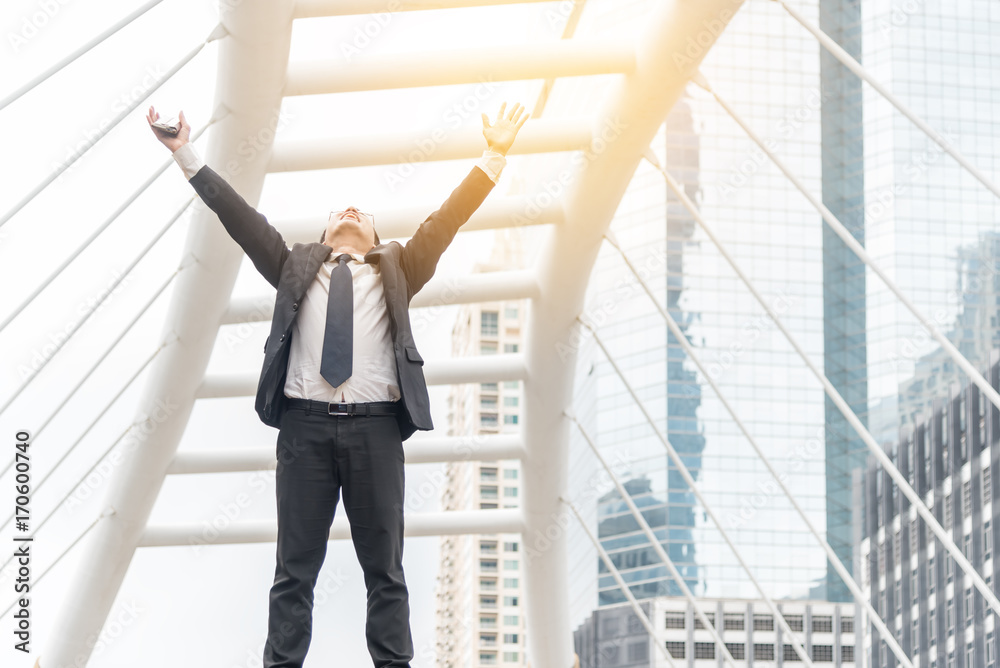 Celebrating success. Low angle view of excited young businessman keeping arms raised and expressing positivity while standing outdoors with office building in the background Celebrating success. Low angle view of excited young businessman keeping arms raised and expressing positivity while standing outdoors with office building in the background