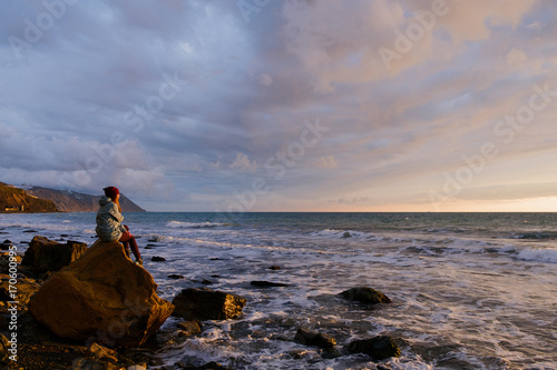 young woman sitting on a rock on the beach and looking at stormy sea and sunset sky