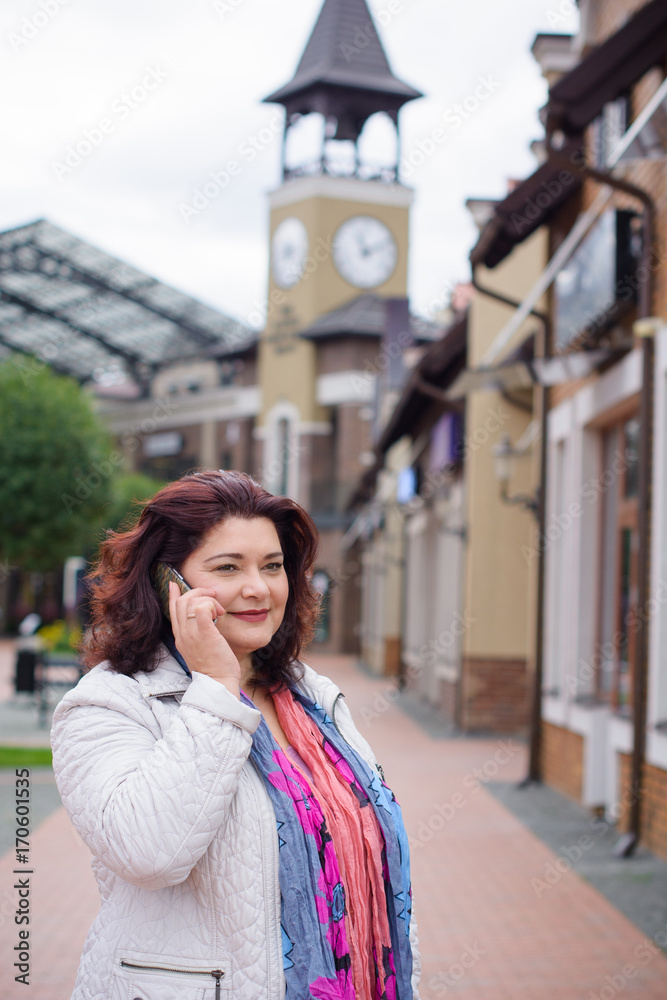 Fototapeta premium Mature cheerful woman talking on a mobile phone in the city and walking among the old part of the city. Good news and good mood