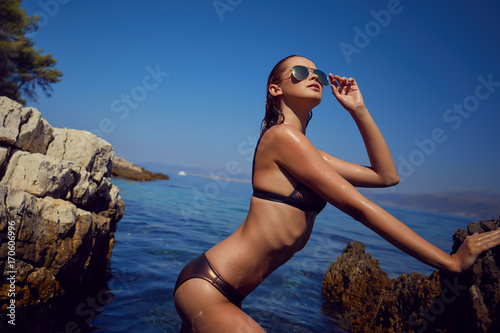 Young adult slim woman in 20s posing on rocks on adriatic sea coast wearing bikini. Toned image.