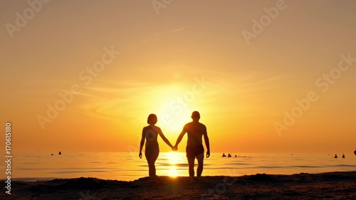 Happy couple walking along a sea beach holding hands on a sunset background